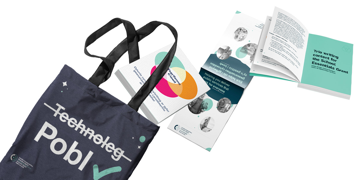 A CDPS tote bag with the Welsh word 'technoleg' crossed out and the Welsh word 'pobl' ticked. Two 'Trio Writing' books and a CDPS brochure are flying out of the bag towards the right.