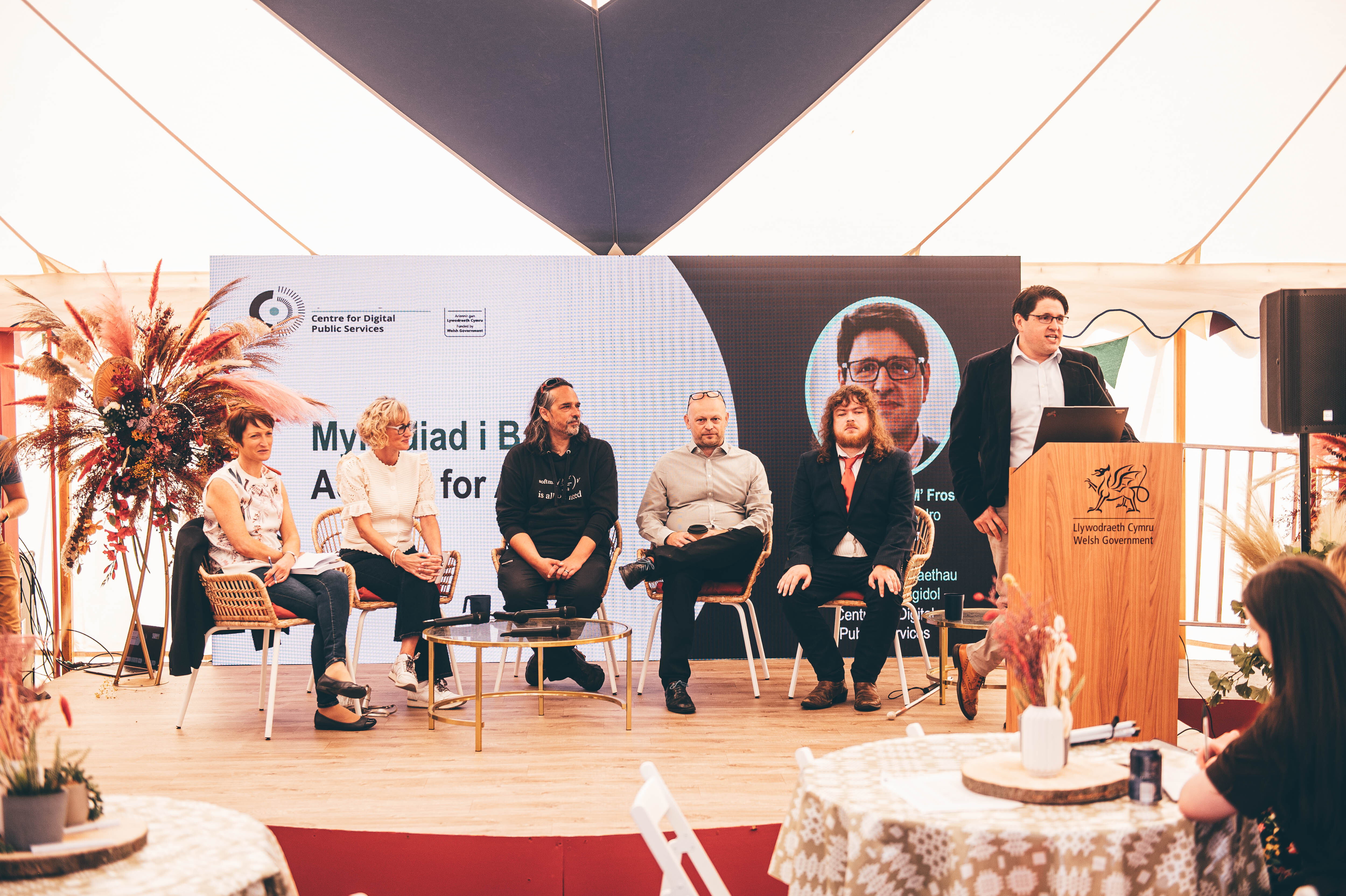 An image of the panel of speakers on stage at the Eisteddfod event. Rhian Bowen Davies, Efa Gruffudd, Stefano Ghazzali, Jeremy Evas and Rob Williams sit in a line on stage, whilst John-Mark Frost stands speaking at the lectern.