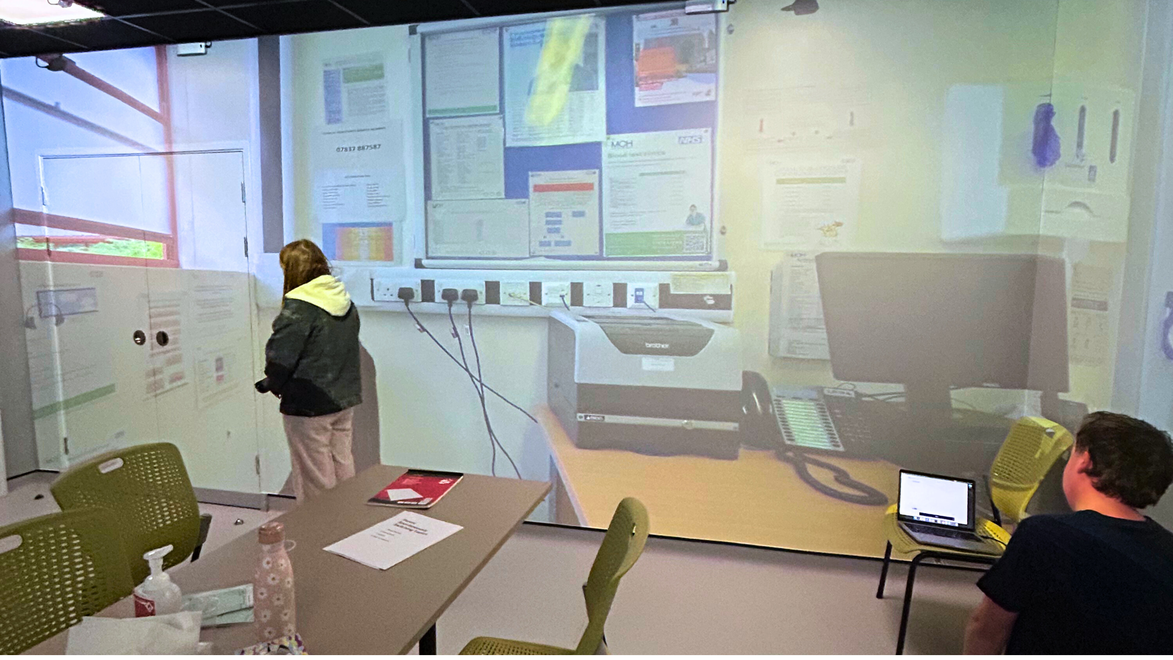 Dave from CDPS and Manon, one of our actors testing the AI scribe tool in Swansea University's simulation suite. The suite has projections on the wall that make it look like a doctor's office. Manon has her back to Dave, to see how the scribe records her speech.
